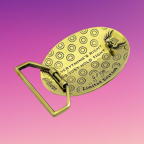 Gold belt buckle with circular patterns and text on a gradient purple and pink background